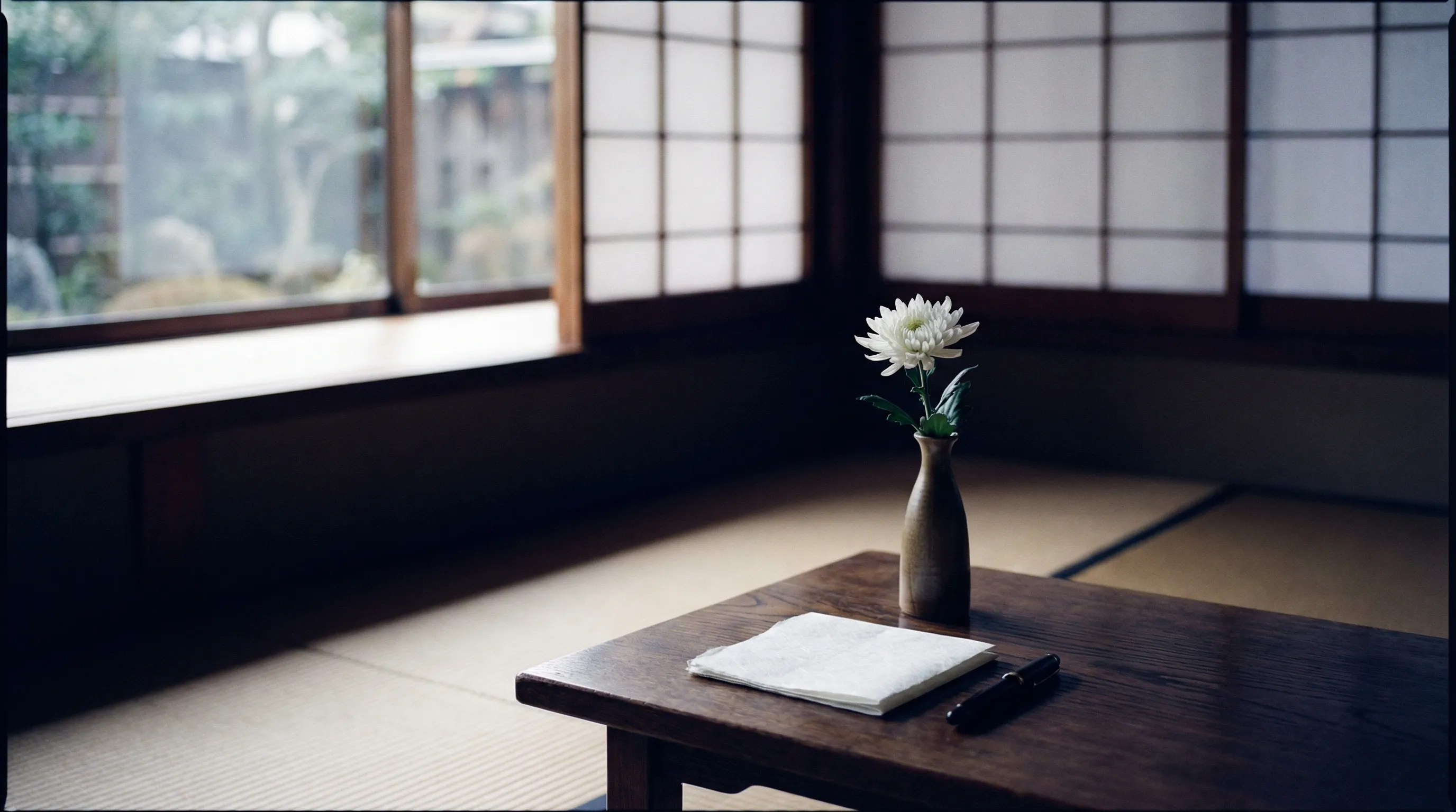障子越しのやわらかな光が差し込む和室に、白い菊の花と筆記具が置かれた静謐な情景
